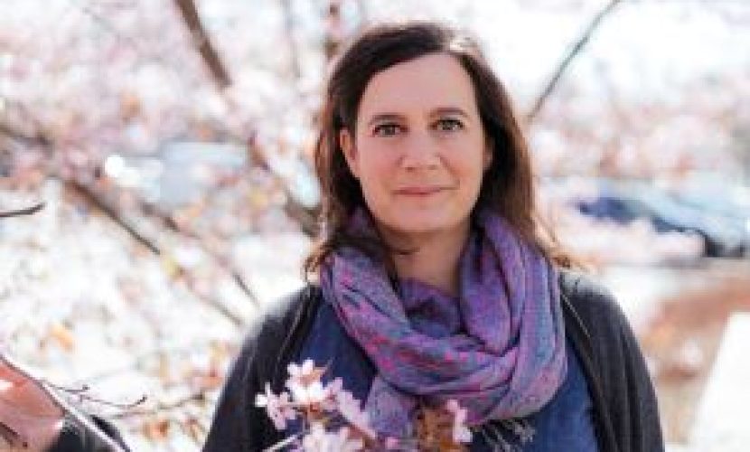 Ilaria tra i ciliegi in fiore. Ilaria ha la pelle chiara, i capelli castani e gli occhi grigi. Indossa una sciarpa viola intorno al collo.