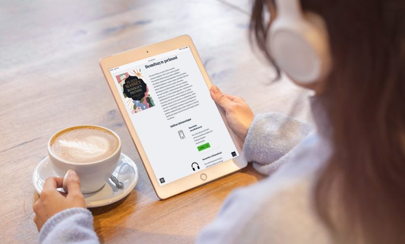Takaviistosta kuvattu pitkätukkainen nuori lukee kuulokkeet päässä tabletilta kirjaa pöydän ääressä. Hän on tarttunut pöydällä olevaan kahvikuppiin.